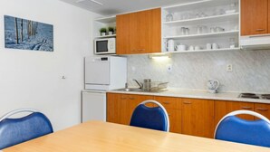 A compact kitchen with wooden cabinets, a microwave, and a refrigerator. There is a dining table with blue chairs and a painting of trees on the wall.