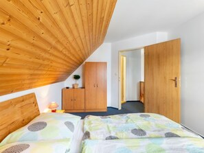 A bedroom with a sloped ceiling, a wooden wardrobe, a bed with patterned bedding, and a small bedside table with a lamp.