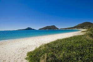 Ein Strand mit weißem Sand, klarem, blauem Wasser und grünen Hügeln im Hintergrund.