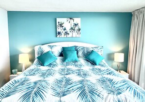 Une chambre à coucher avec un lit orné d’un motif de feuilles de palmier, une tête de lit, deux lampes de chevet et un tableau représentant des palmiers accroché au mur.