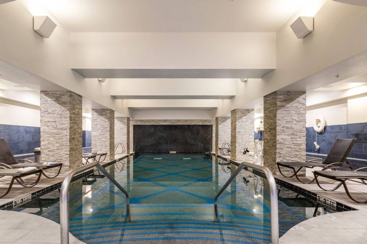 A modern indoor swimming pool with clear lanes, lounge chairs, and a stone wall backdrop.