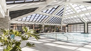 An indoor swimming pool with a high ceiling and skylights, surrounded by seating areas and potted plants.