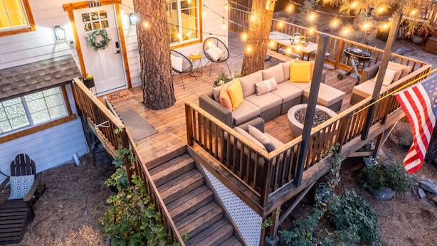 A wooden deck with a sofa and chairs, surrounded by trees and string lights. - Cabin Near Ski Resort, EV Charge, Kid/Pet Friendly, Fire Pit, Playhouse (Wrightwood)