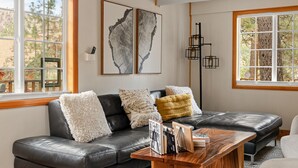 A modern living room with a black leather sectional sofa, a wooden coffee table, and a large window with a view of trees. - Cabin Near Ski Resort, EV Charge, Kid/Pet Friendly, Fire Pit, Playhouse (Wrightwood)