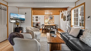 A cozy living room with a fireplace, a dining area, and a large window. - Cabin Near Ski Resort, EV Charge, Kid/Pet Friendly, Fire Pit, Playhouse (Wrightwood)