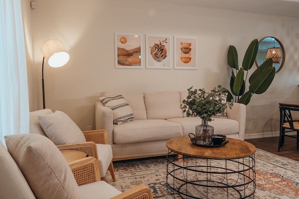 A living room with a sofa, two armchairs, a coffee table, and a large plant. There are three framed artworks on the wall and a floor lamp.