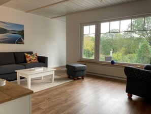 A living room with a sofa, ottoman, coffee table, and a large window with a view of trees.