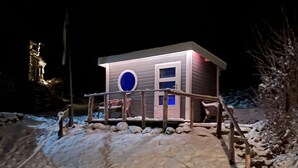 A small, single-story building with a circular window and a door, surrounded by a wooden deck and snow.