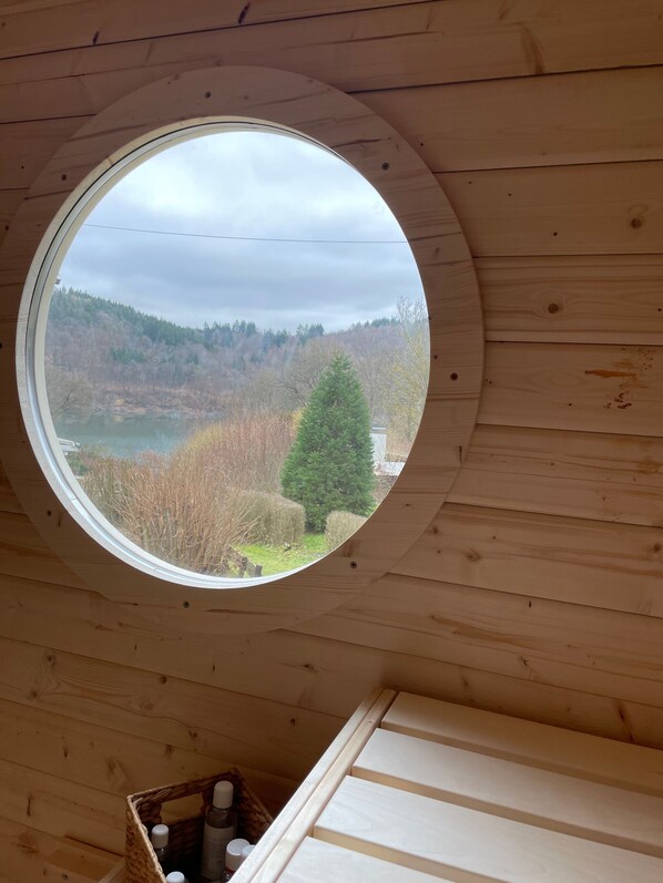 A circular window in a wooden cabin with a view of a forested landscape and a lake.