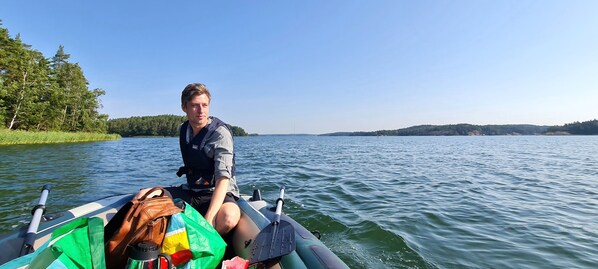 Une personne se trouve dans un petit bateau, avec un sac et une pagaie, entourée d’une eau calme et d’arbres.