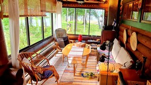 A cozy cabin interior with a large window overlooking a lush garden, a wooden bench, a wicker chair, and a small table with a lamp. - A Real Home - sea View Close to City Center (Pargas)