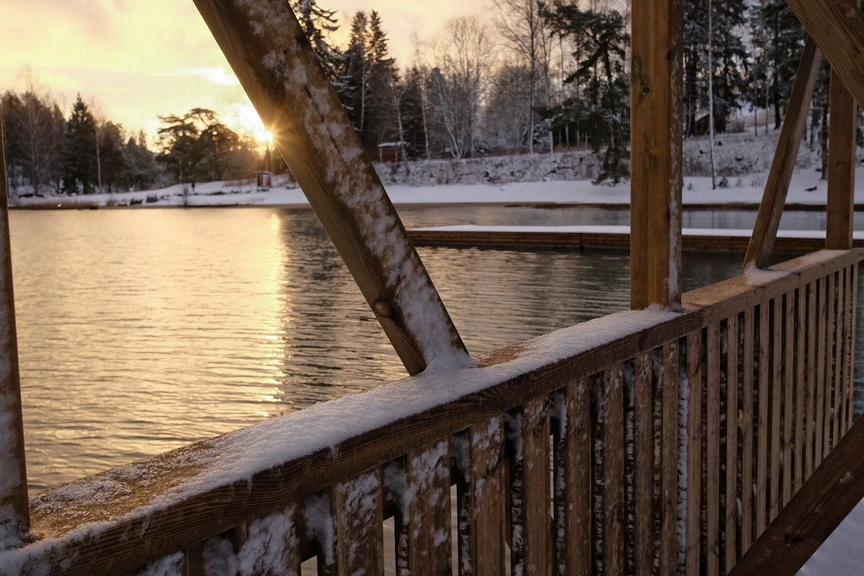 A wooden deck with snow accumulation, overlooking a calm body of water and a forested area during sunset.