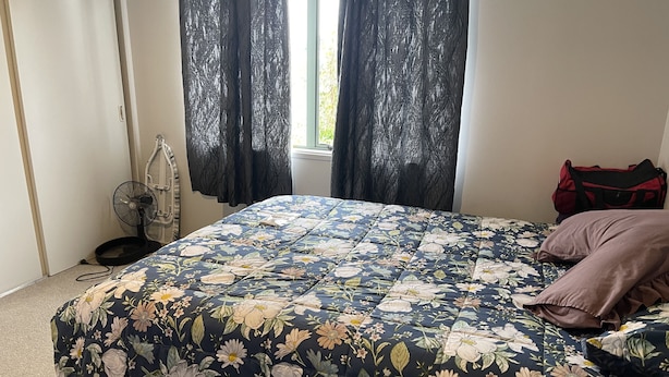 Een slaapkamer met een bloemenbedekking, een staande ventilator, een raam met gordijnen en een rode tas op het bed.