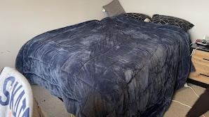 Een slaapkamer met een groot bed bekleed met een donkerblauwe sprei, een houten nachtkastje met een lamp, en een klein lade.
