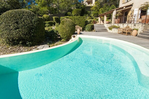 Una piscina curvada con agua azul cristalina, rodeada de follaje cuidado y un muro de piedra.