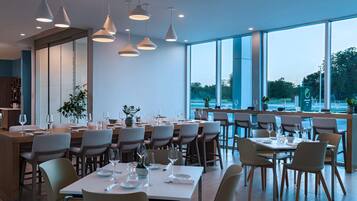 A modern restaurant with multiple dining tables and chairs, large windows offering a view of the outdoors, and pendant lighting.