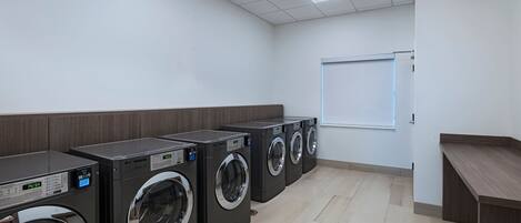 A row of front-loading washing machines in a clean, tiled laundry room with a whiteboard on the wall.