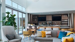 Una sala de estar moderna con un sofá gris, sillones y una mesa de centro. Se observa un área de bar con estantes llenos de botellas y vasos al fondo.