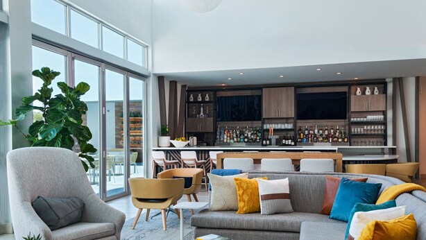 Un salon moderne avec un canapé gris, des fauteuils et une table basse. On aperçoit à l’arrière-plan un coin bar avec des étagères remplies de bouteilles et de verres.