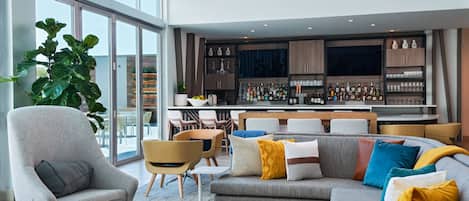 A modern living room with a grey sofa, armchairs, and a coffee table. A bar area with shelves of bottles and glasses is visible in the background.