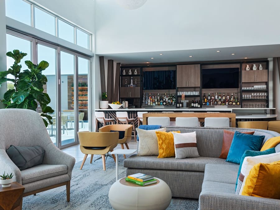 A modern living room with a grey sofa, armchairs, and a coffee table. A bar area with shelves of bottles and glasses is visible in the background.