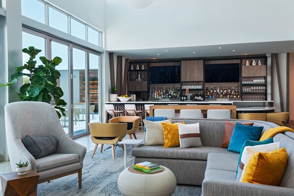 Una sala de estar moderna con un sofá gris, sillones y una mesa de centro. Se observa un área de bar con estantes llenos de botellas y vasos al fondo.