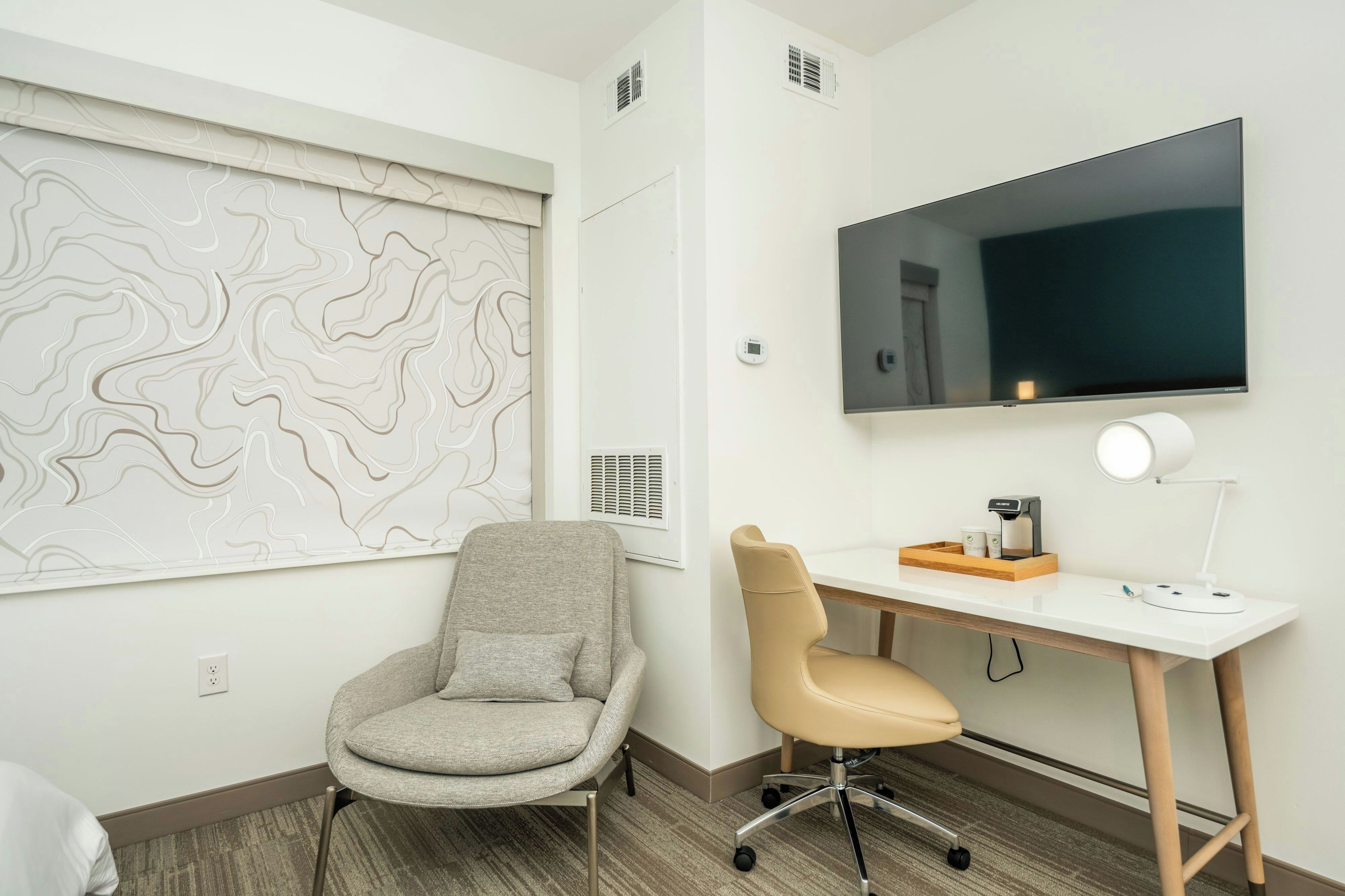 A modern office space with a desk, two chairs, a large flat-screen TV, a coffee machine, and a wall-mounted air conditioning unit.