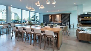 Un comedor moderno con una larga mesa de madera y sillas blancas, ventanas amplias y una encimera de cocina con un menú en una pizarra.