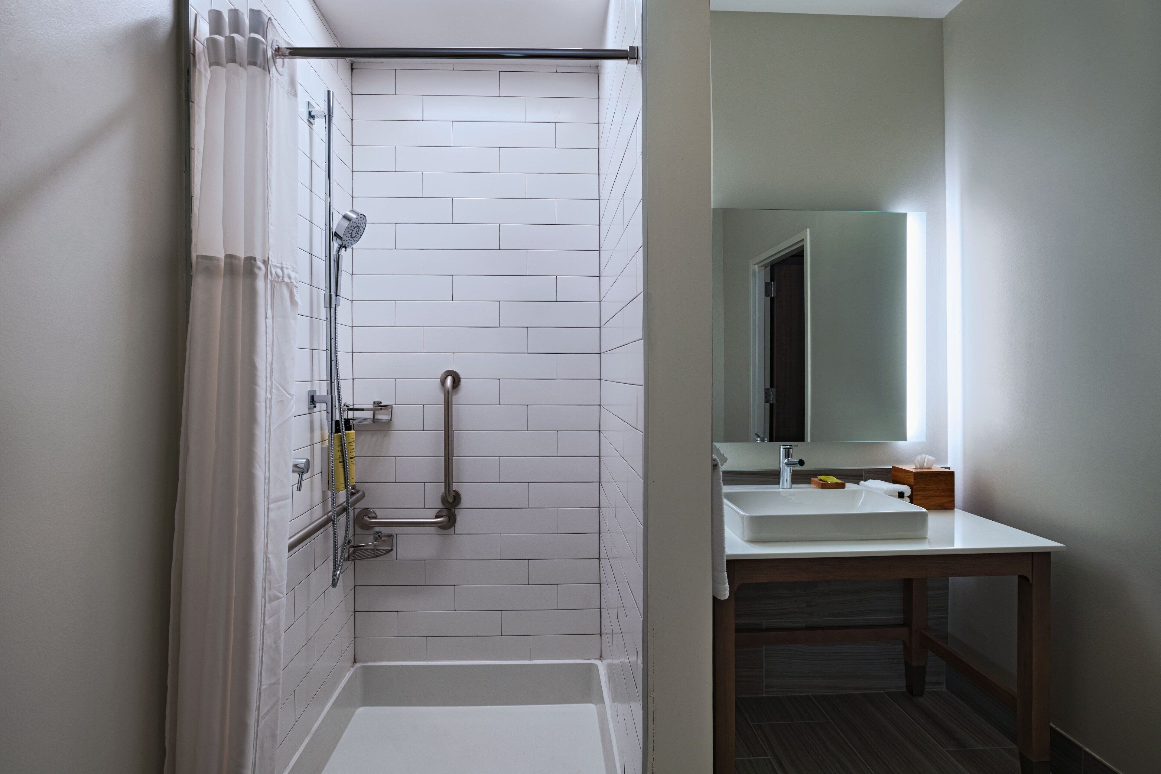 A modern bathroom with white tiled walls, a shower with a glass door, a white sink, and a wooden vanity.