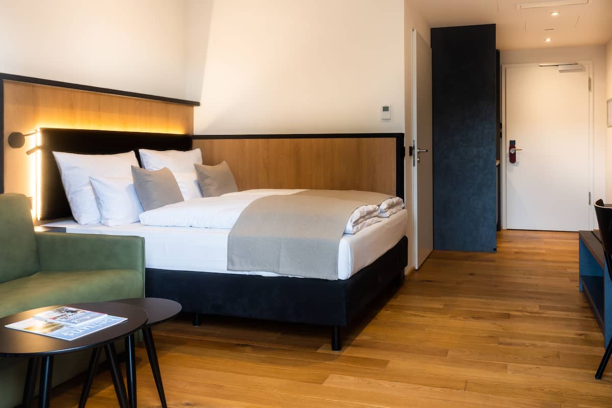 Une chambre d’hôtel moderne avec un lit, un fauteuil vert, une petite table et un plancher en bois.