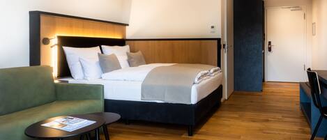 Una habitación de hotel moderna con una cama, un sillón verde, una mesita pequeña y suelo de madera.