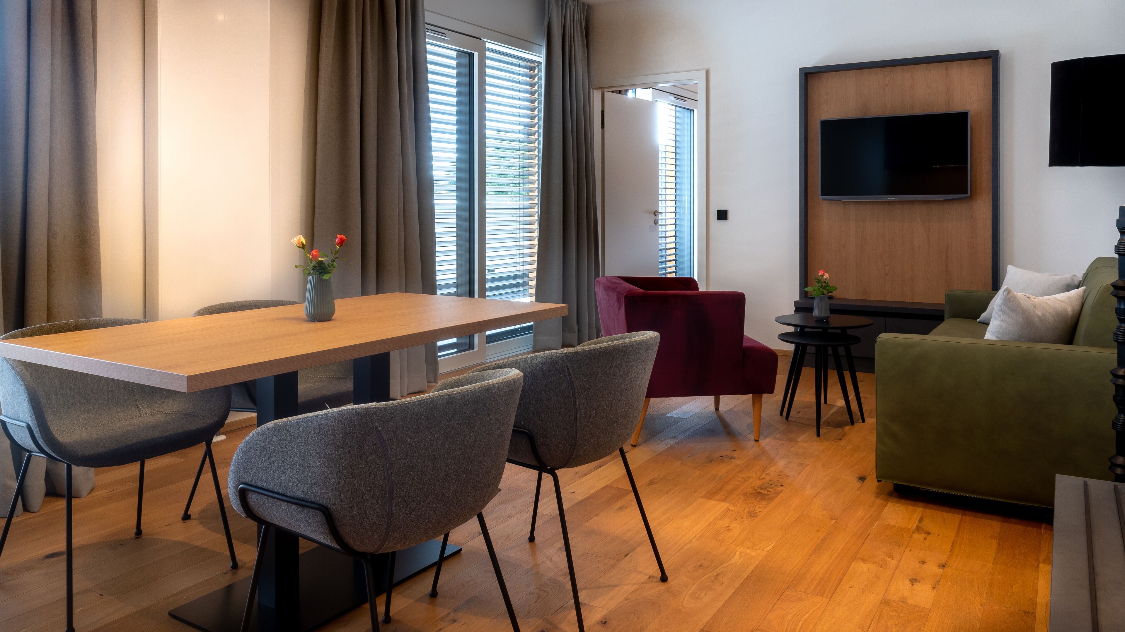A modern living room with a wooden dining table, grey upholstered chairs, a red armchair, a green sofa, and a flat-screen TV mounted on the wall.