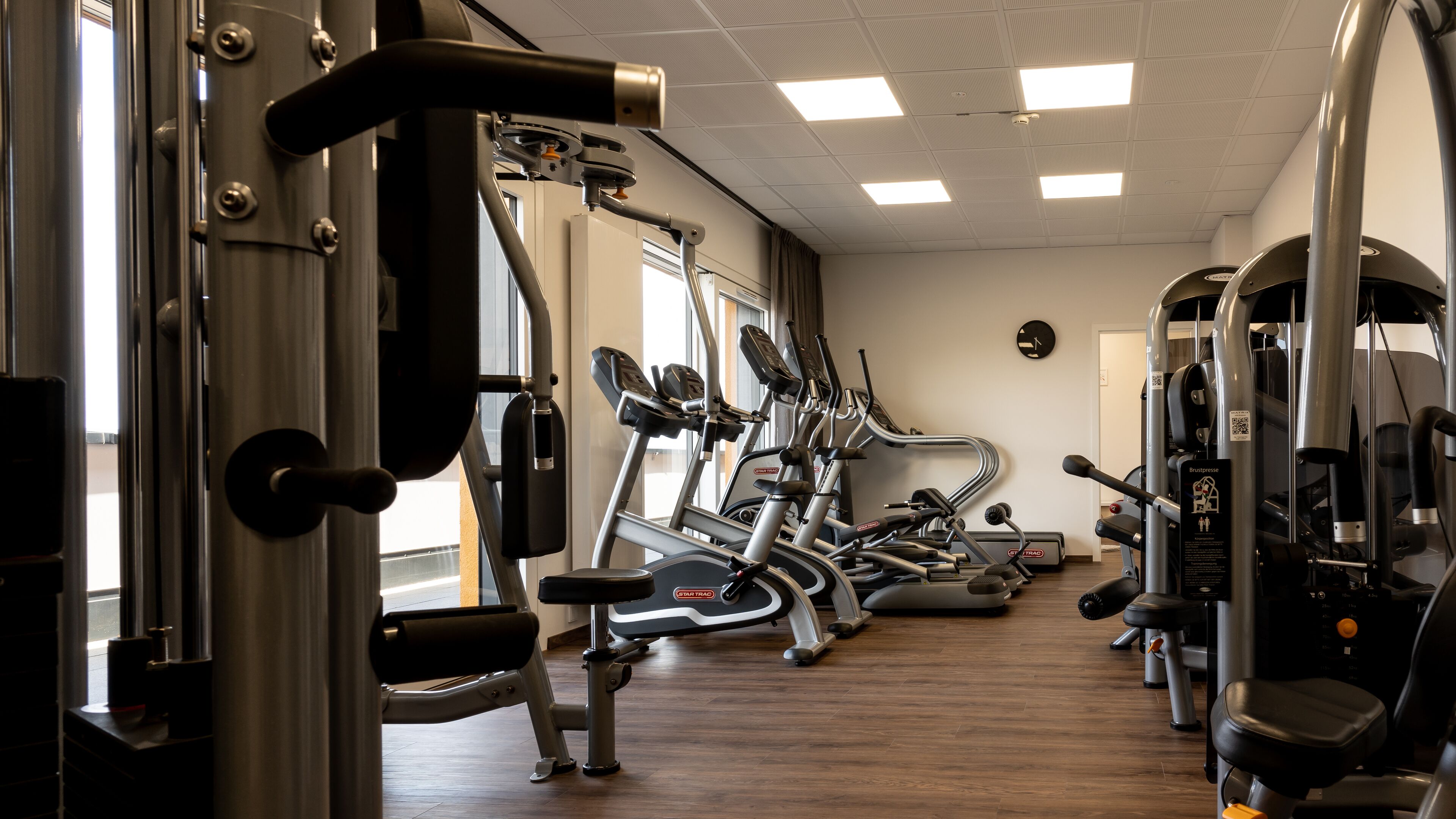 Ein Fitnessraum mit verschiedenen Trainingsgeräten, einer Uhr an der Wand und einem Fenster mit Vorhängen.