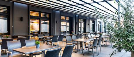 An outdoor cafe with tables and chairs, a pergola with lights, and a view of the street.