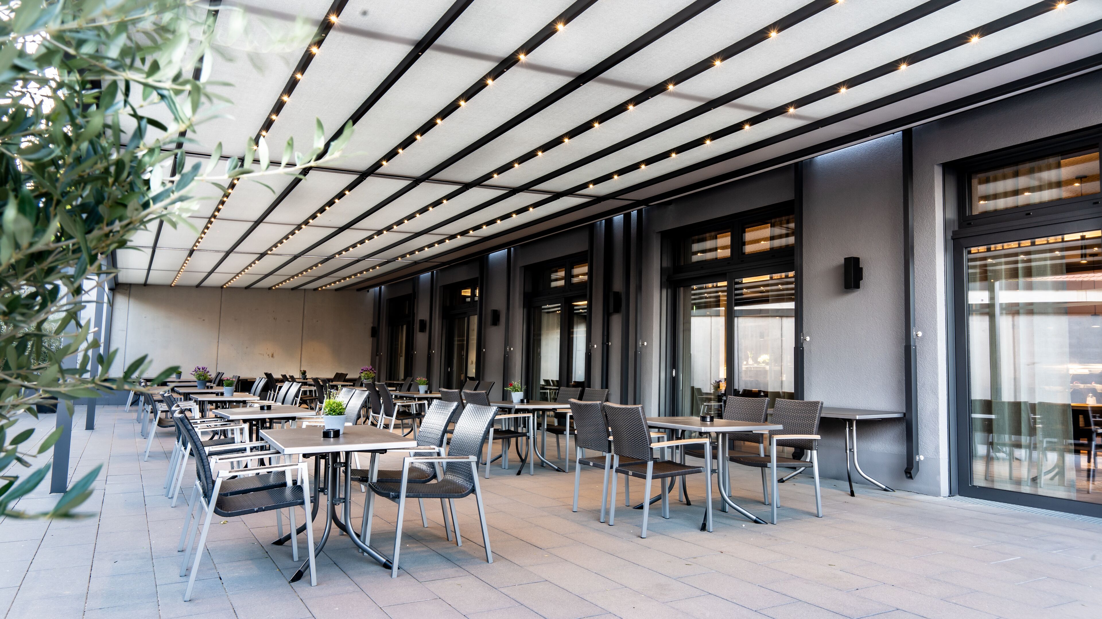 An outdoor seating area with tables and chairs, a ceiling with lights, and large windows.