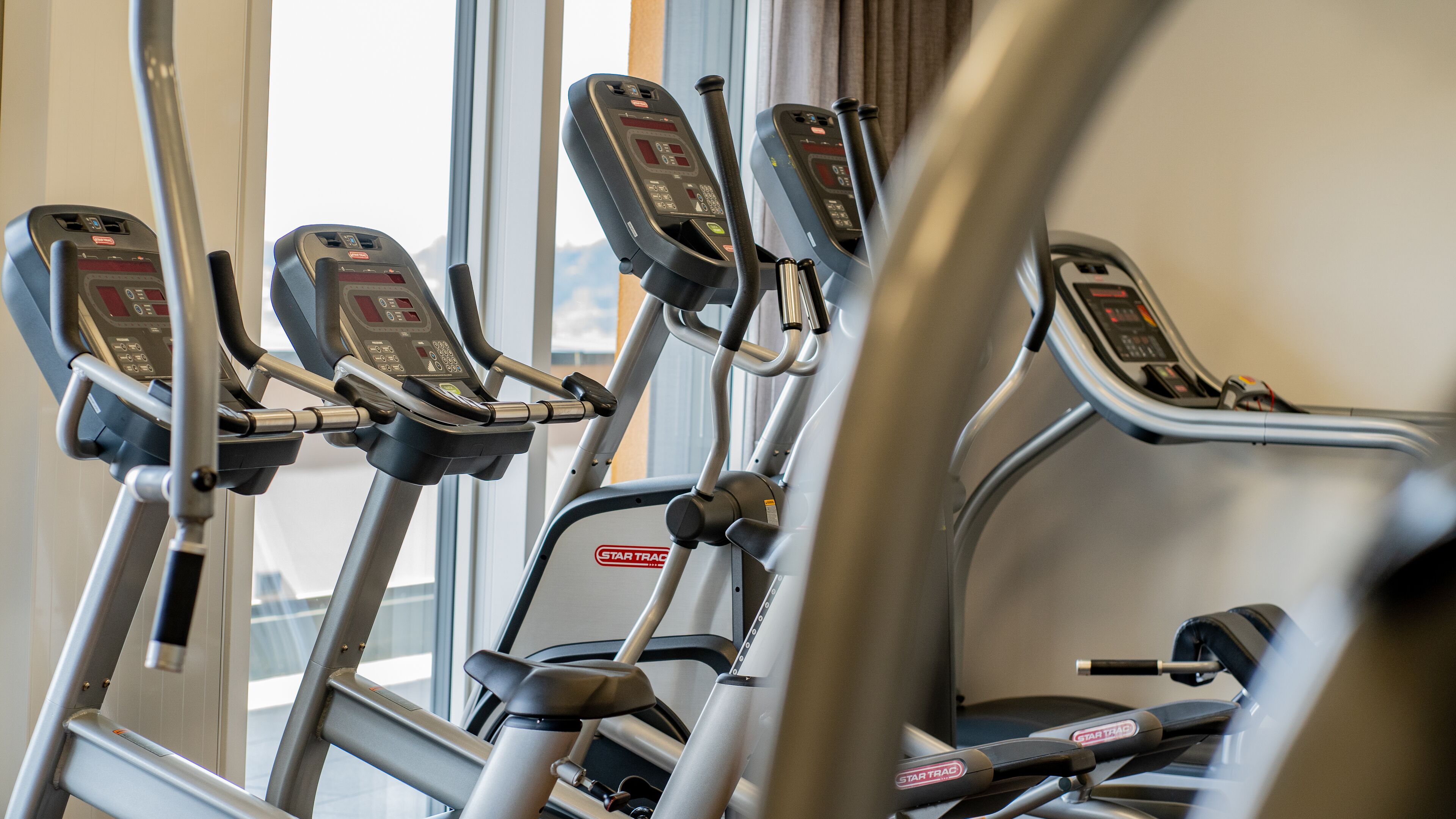 A row of stationary exercise bikes with digital displays, positioned in a gym with large windows.