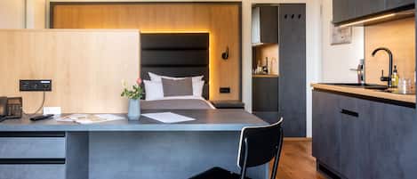 Habitación de hotel moderna con una isla central, una cama con cabecera, una pequeña cocina y un baño visible a través de una puerta abierta.