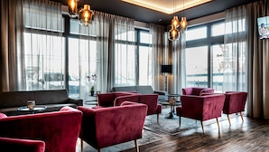 A modern hotel lobby with large windows, red upholstered chairs, a grey sofa, and a small table with a lamp. - Burgau Hotel Sonnenhof (Burgau)
