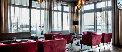 Un hall d’hôtel moderne avec de grandes fenêtres, des fauteuils à revêtement rouge, un canapé gris et une petite table avec une lampe.