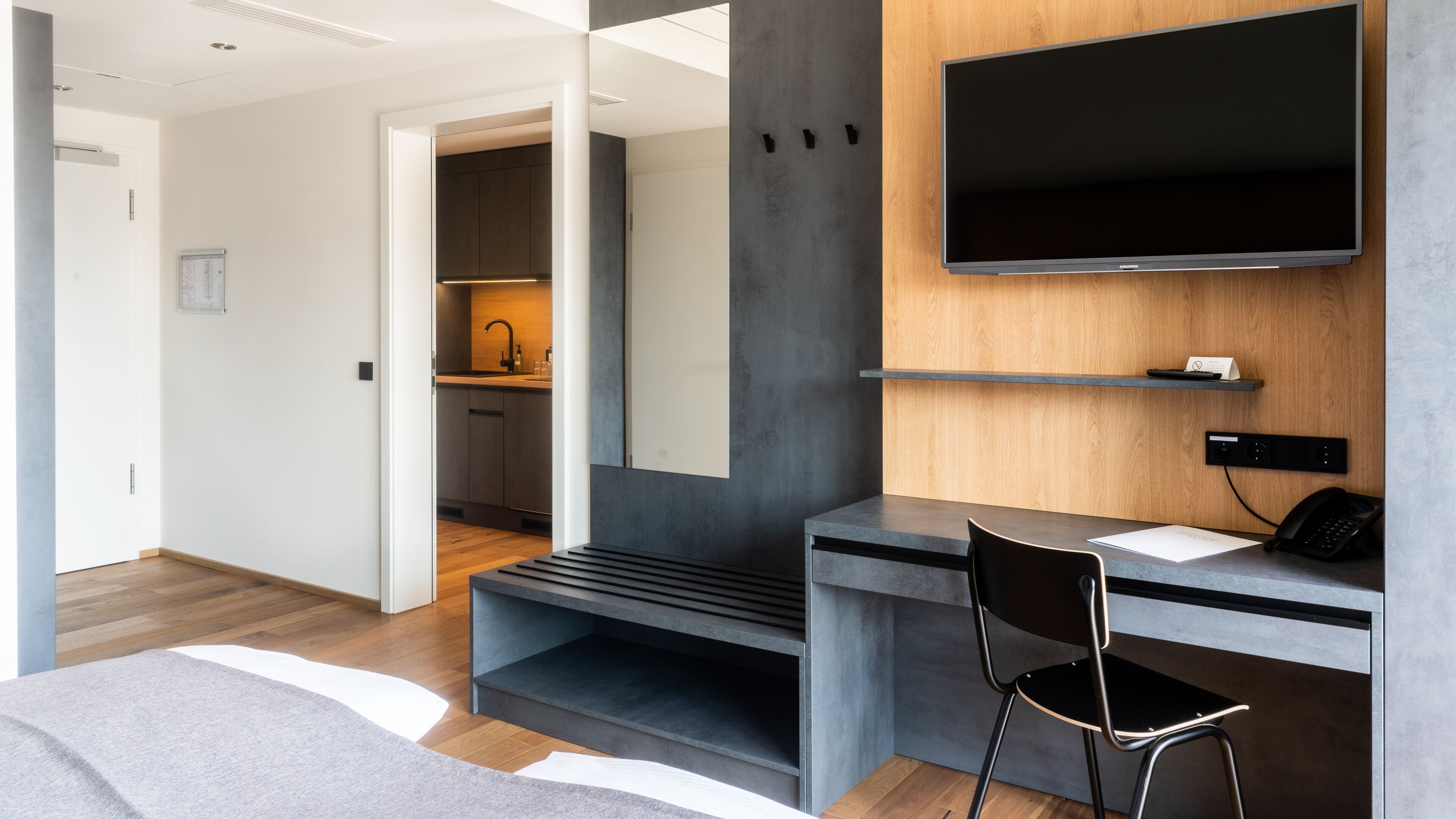 A modern hotel room with a large flat-screen TV mounted on a wooden wall, a desk with a chair, and a visible kitchen area with a sink and cabinets.