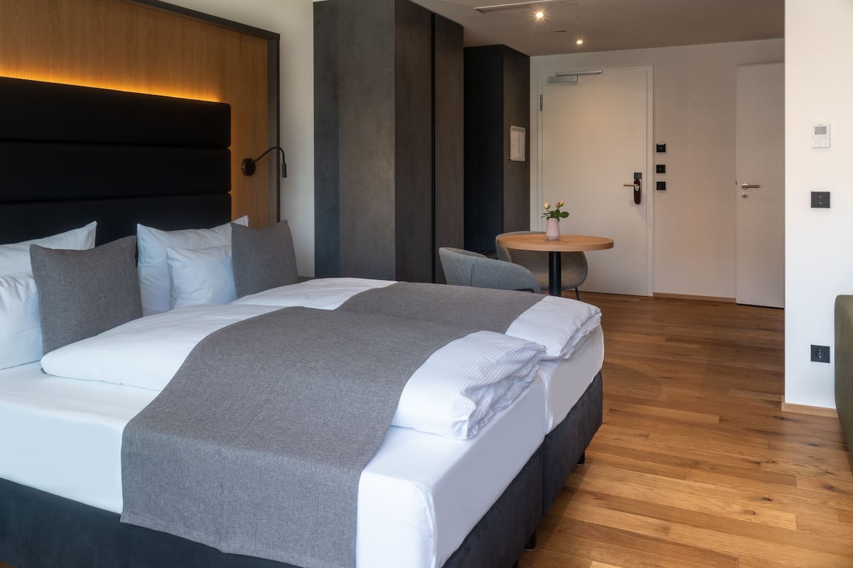 Une chambre d’hôtel moderne avec un grand lit, des tables de chevet, une table ronde ornée d’un vase et un plancher en bois.