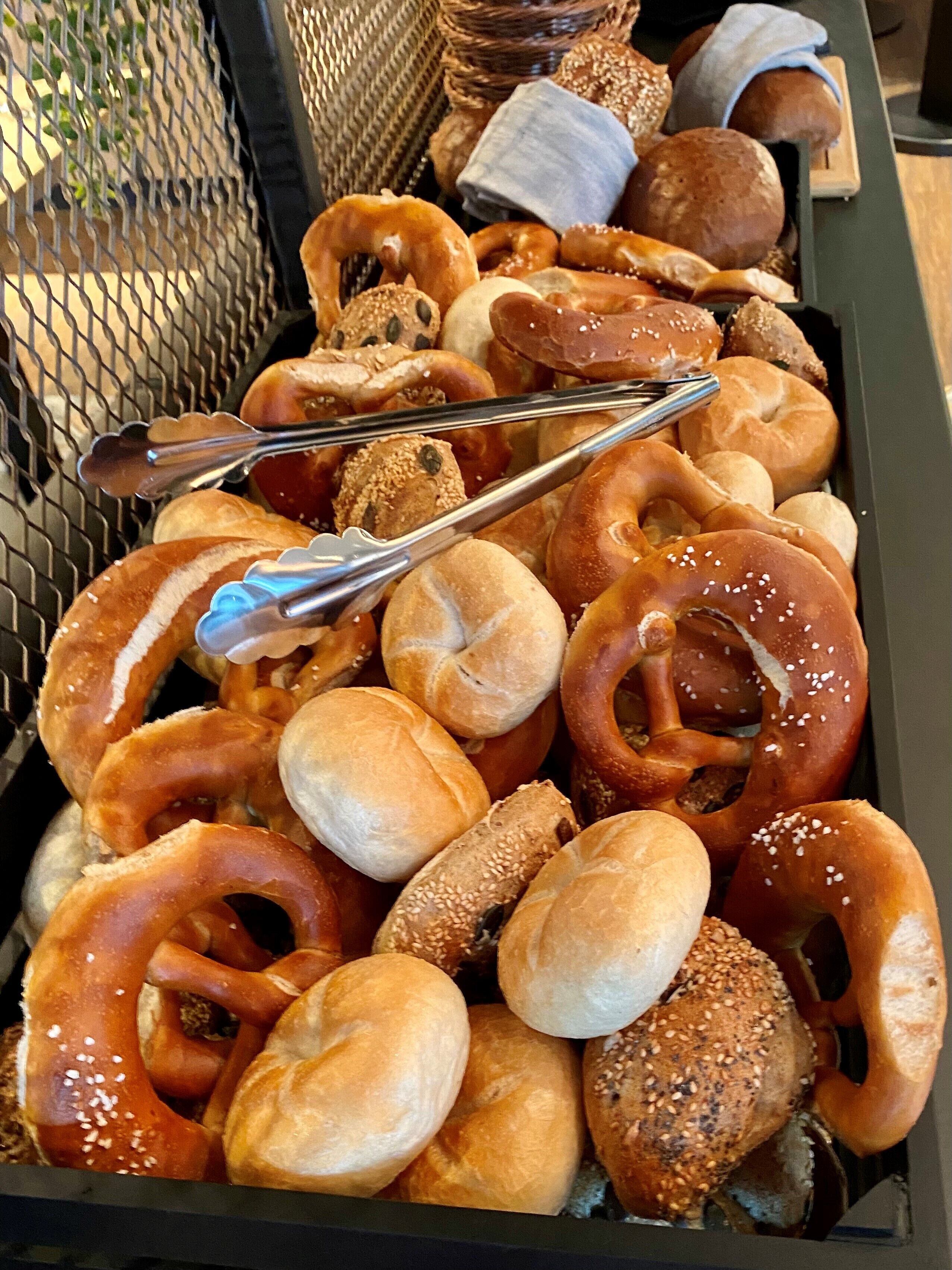 Una bandeja con varios productos horneados, incluyendo pretzels y panecillos, con unas pinzas encima.