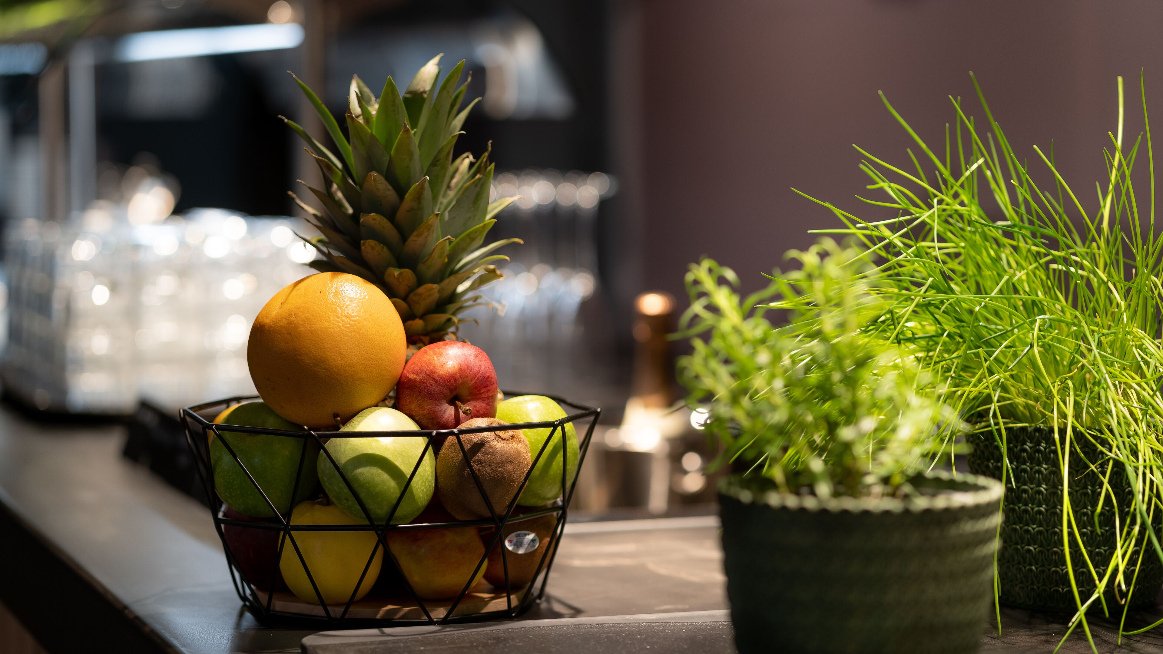 A fruit bowl with apples, oranges, and a pineapple on a table next to a potted plant.
