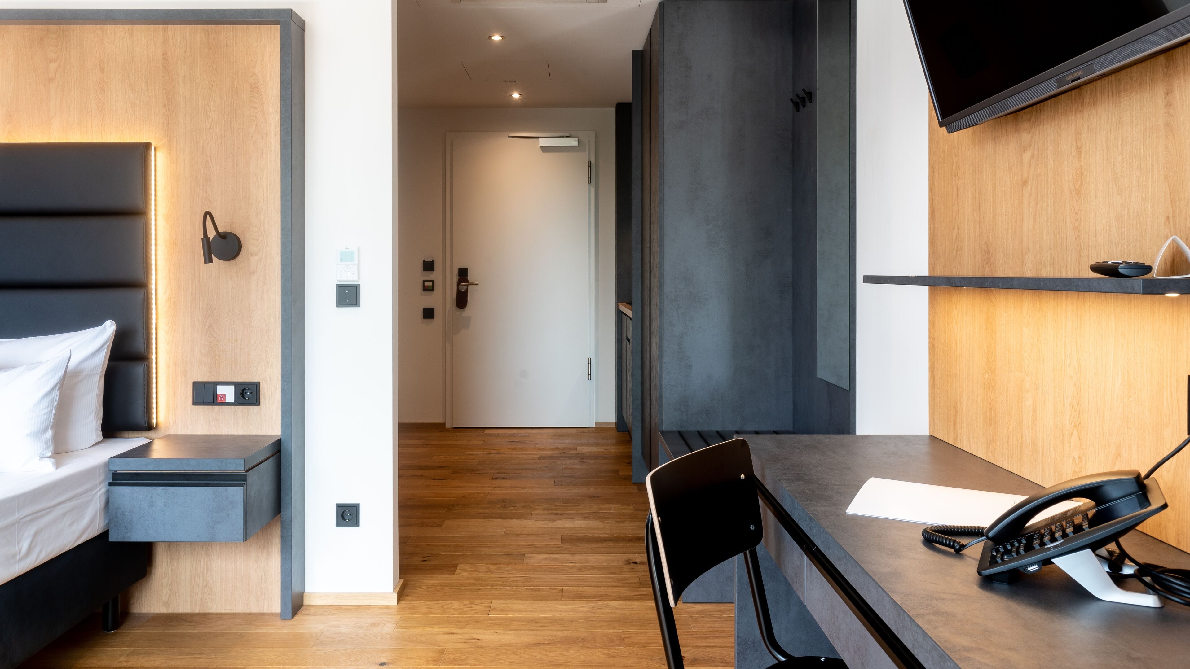 A modern hotel room with a bed, bedside table, desk, chair, and a telephone.