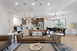 A modern living room with a brown leather sofa, a dining area with a glass table, and a kitchen with stainless steel appliances.