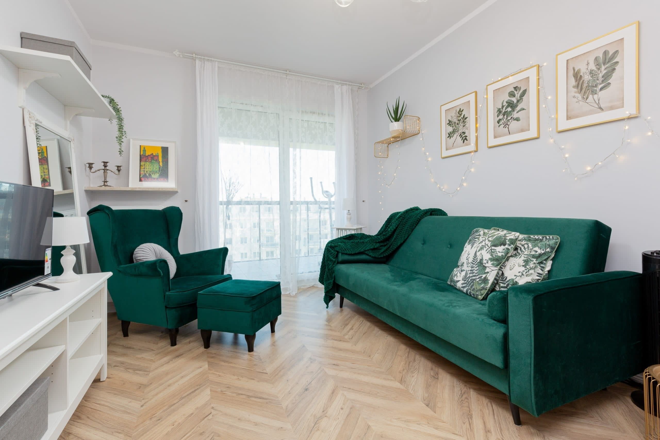 A living room with a green sofa, matching ottoman, and a TV on a white stand. There are framed botanical prints on the wall and a plant in a basket.