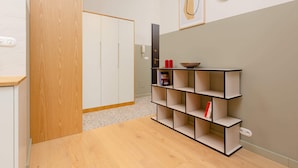 A modern room with a wooden wardrobe, a multi-tiered black shelving unit, and a plain wall with a framed abstract artwork.