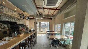A spacious cafe with a long counter, bar stools, and tables. There are shelves with bottles and glasses, and a view of the street through large windows.