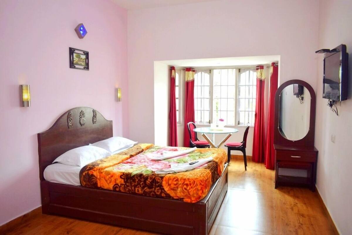 Een slaapkamer met een houten bed, een klein tafeltje met stoelen, een spiegel en een raam met gordijnen.