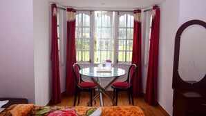 Een kamer met een ronde glazen tafel en twee stoelen, een bed met een kleurrijke deken, en een groot raam met rode gordijnen.